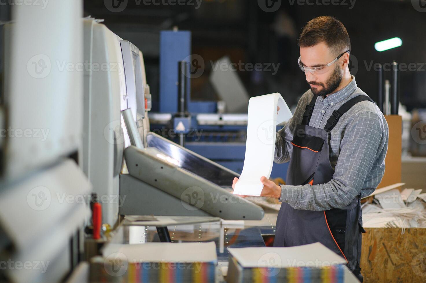 Printing house interior with modern offset print machine and operator in working uniform checking quality and controlling process of print photo