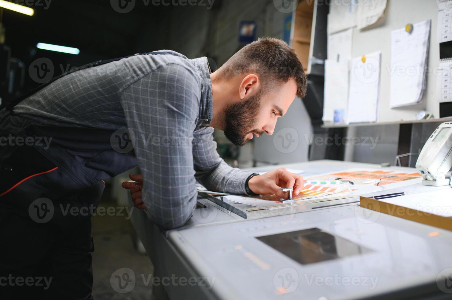 Printing house interior with modern offset print machine and operator in working uniform checking quality and controlling process of print photo