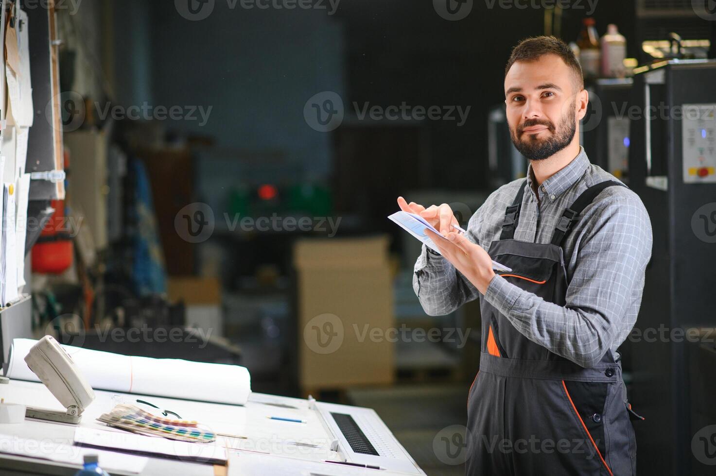 Printing house interior with modern offset print machine and operator in working uniform checking quality and controlling process of print photo