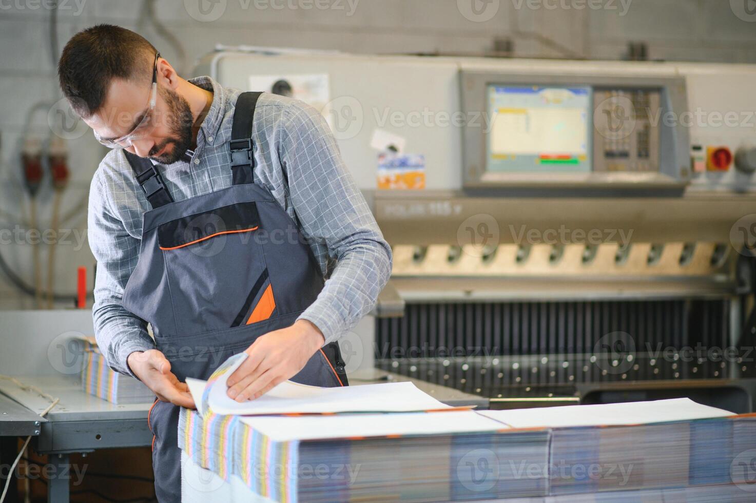 Printing house interior with modern offset print machine and operator in working uniform checking quality and controlling process of print photo