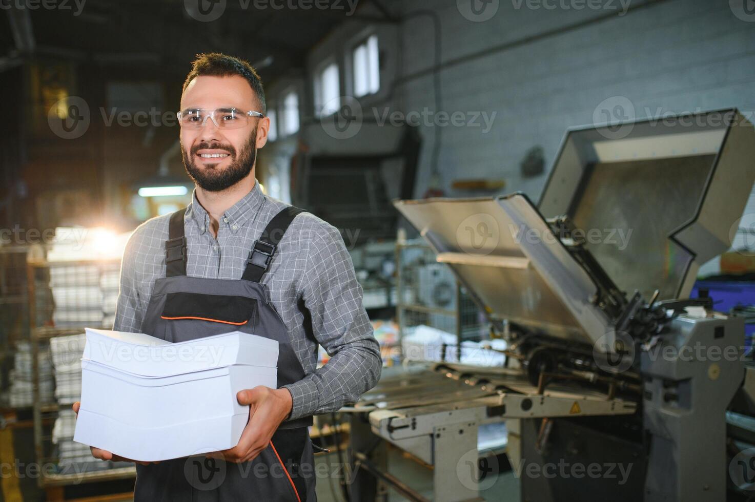 Printing house interior with modern offset print machine and operator in working uniform checking quality and controlling process of print photo