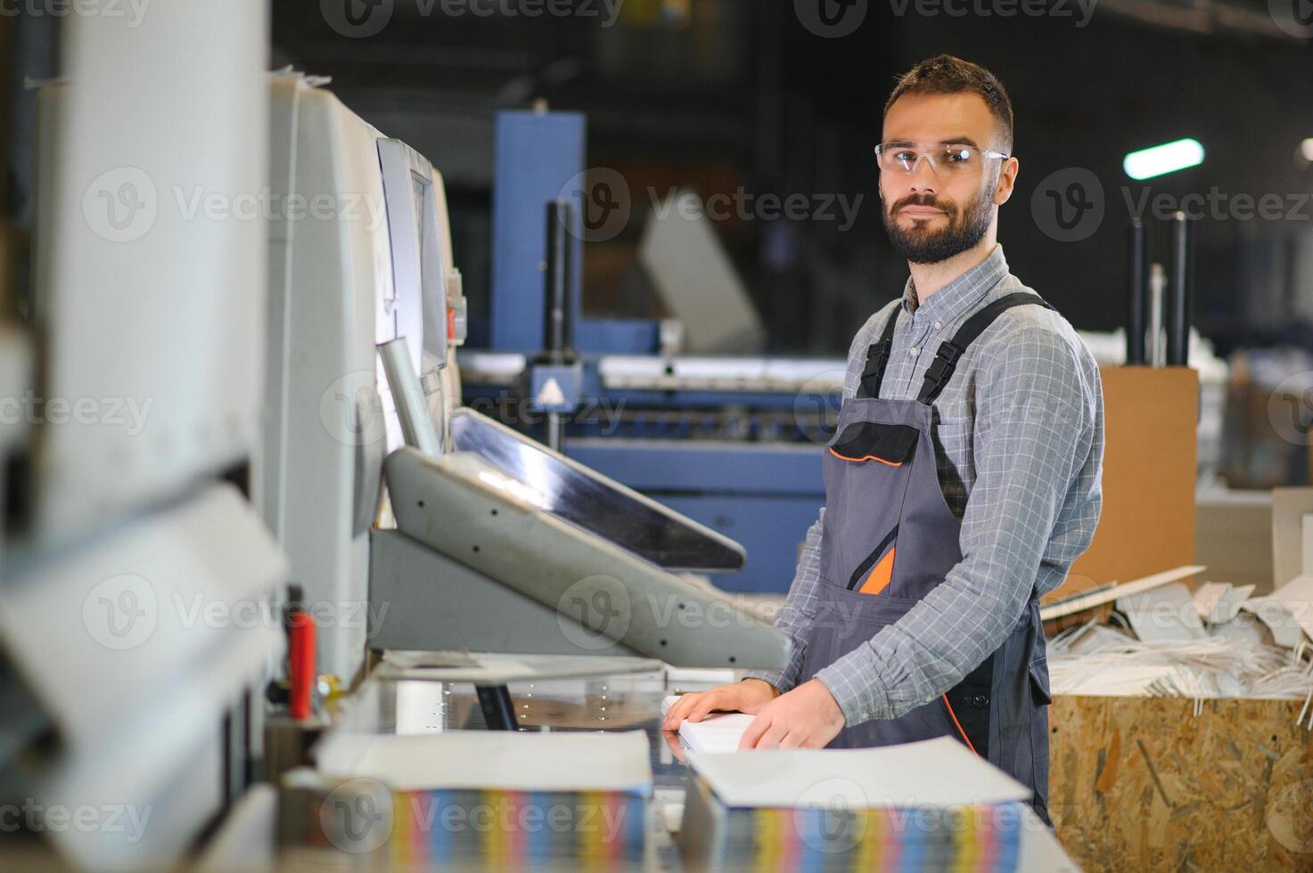 Printing house interior with modern offset print machine and operator in working uniform checking quality and controlling process of print photo