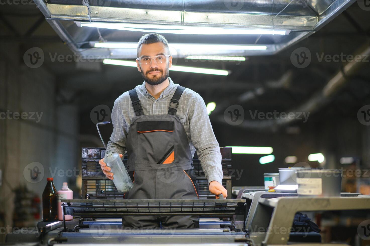 Printing house interior with modern offset print machine and operator in working uniform checking quality and controlling process of print photo