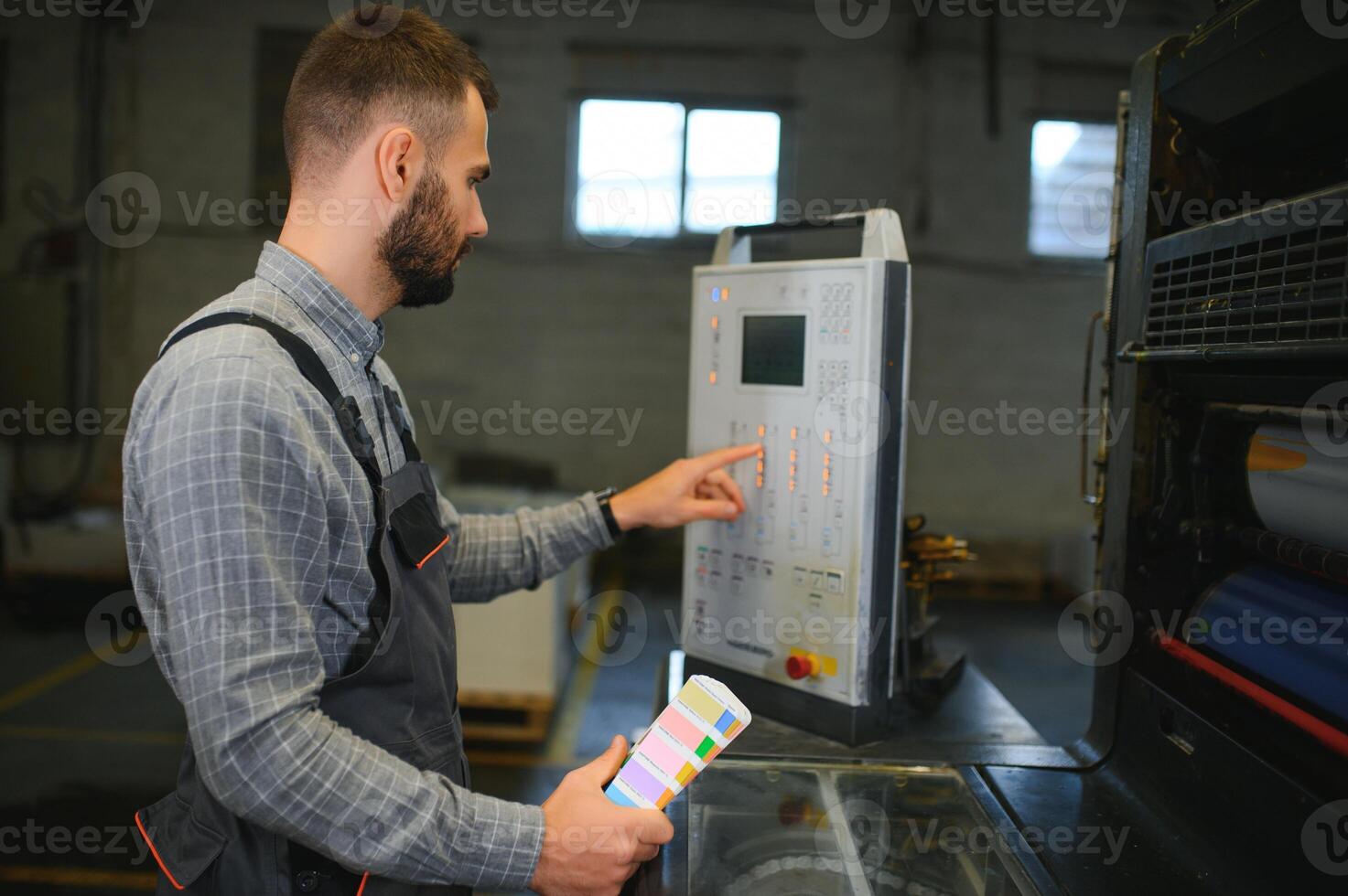 Printing house interior with modern offset print machine and operator in working uniform checking quality and controlling process of print photo