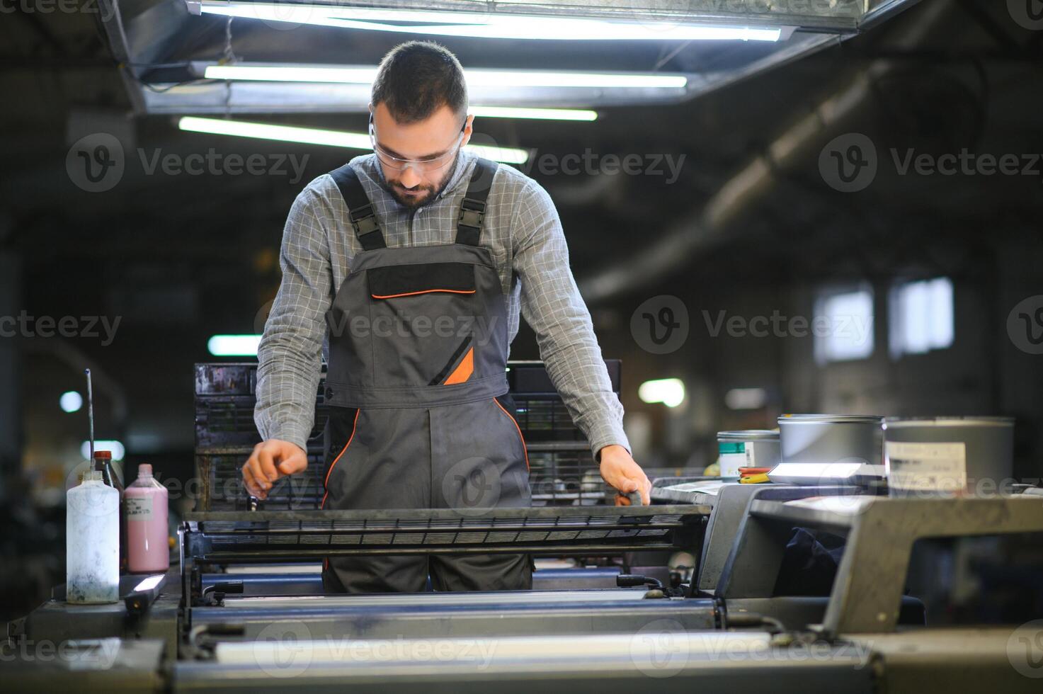 Printing house interior with modern offset print machine and operator in working uniform checking quality and controlling process of print photo