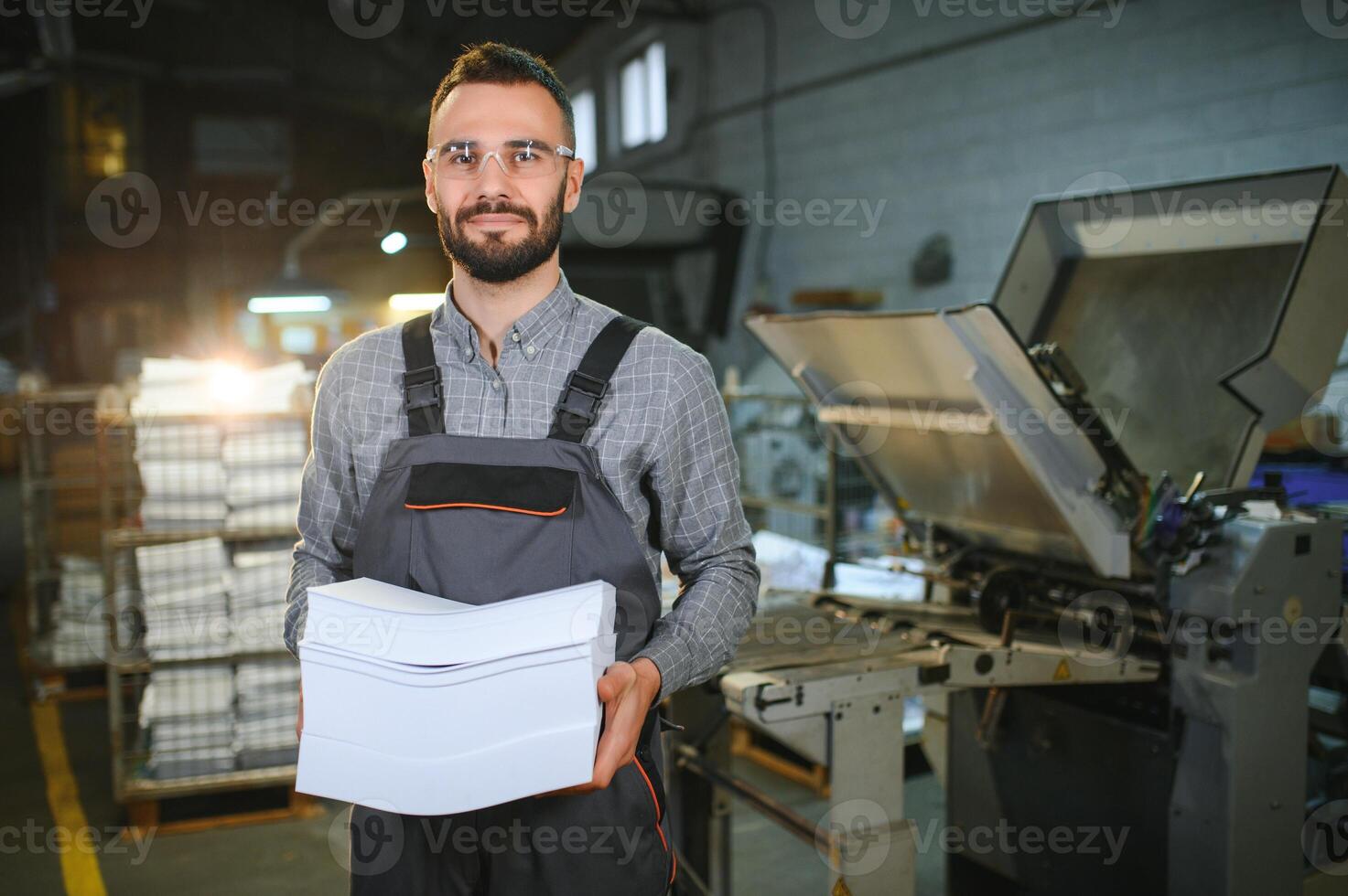 Printing house interior with modern offset print machine and operator in working uniform checking quality and controlling process of print photo