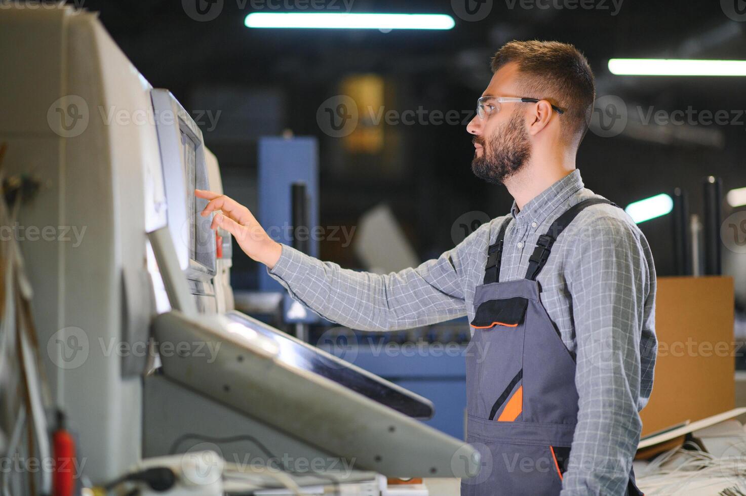 Printing house interior with modern offset print machine and operator in working uniform checking quality and controlling process of print photo