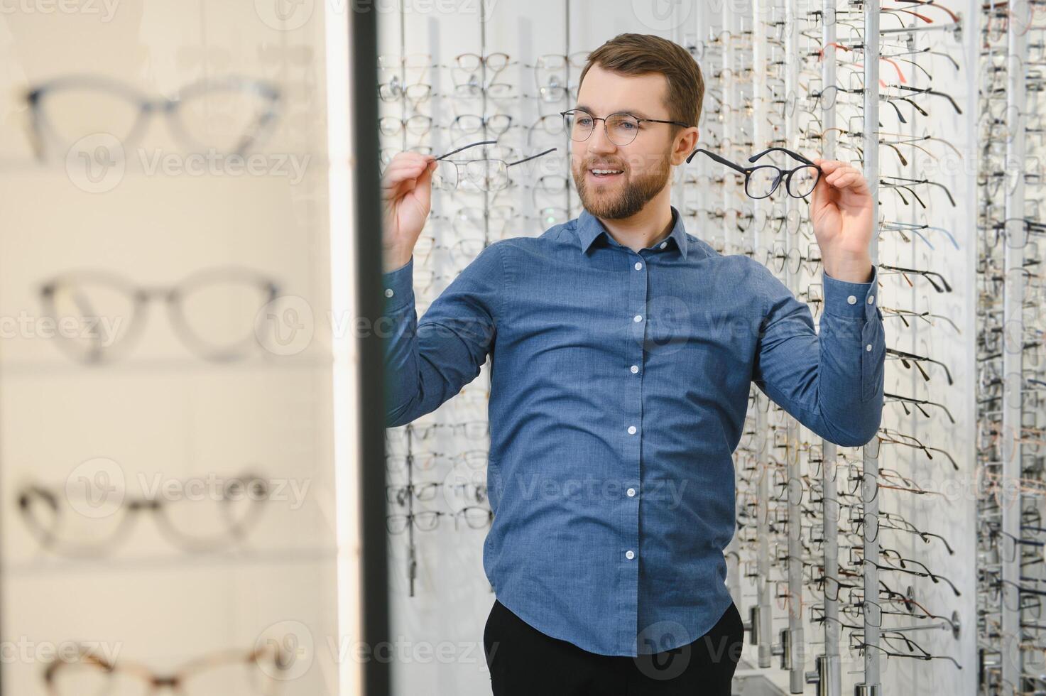 In Optics Shop. Portrait of male client holding and wearing different spectacles, choosing and trying on new glasses at optical store. Man picking frame for vision correction, closeup. photo