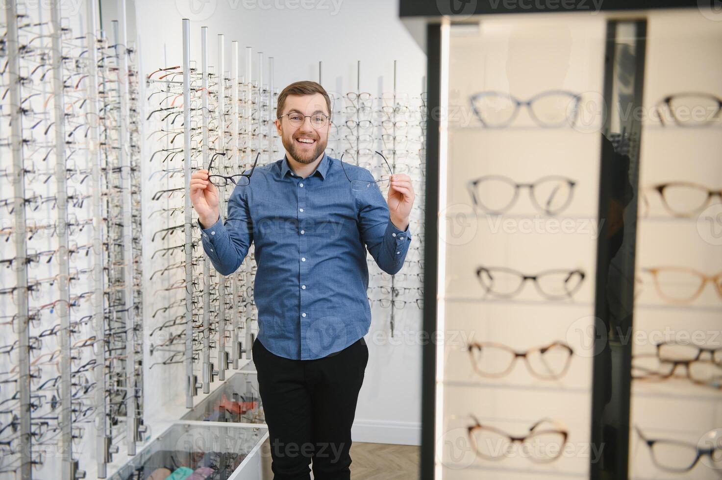 en óptica tienda. retrato de masculino cliente participación y vistiendo diferente gafas, elegir y molesto en nuevo lentes a óptico almacenar. hombre cosecha marco para visión corrección, de cerca. foto