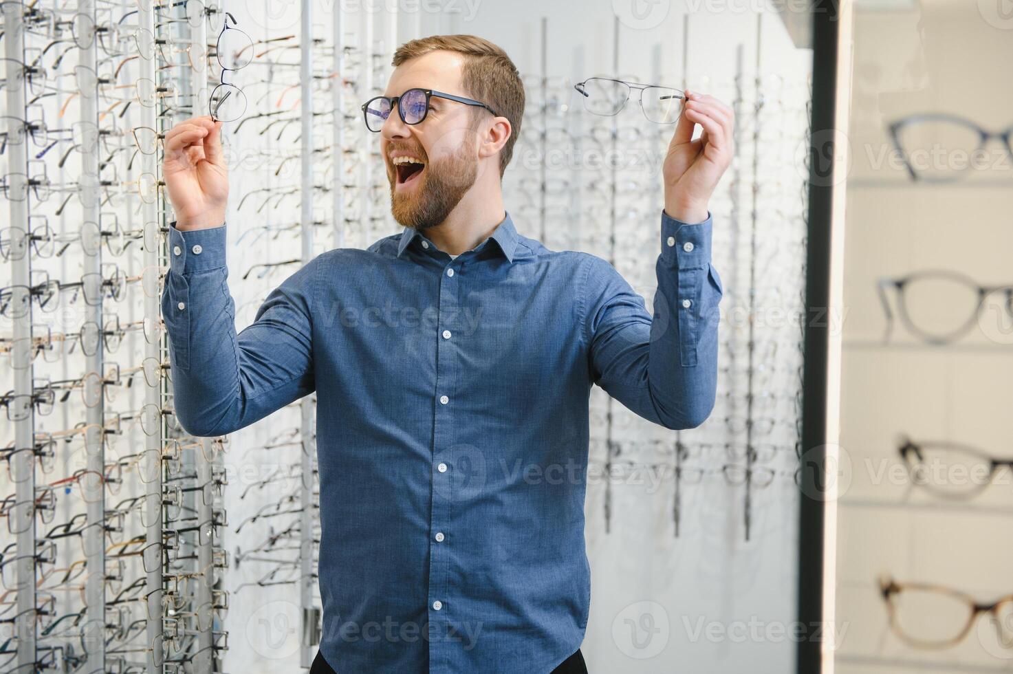 Satisfied Customer. View of happy young male client wearing new glasses, standing near rack and showcase with eyewear. Smiling man trying on spectacles photo