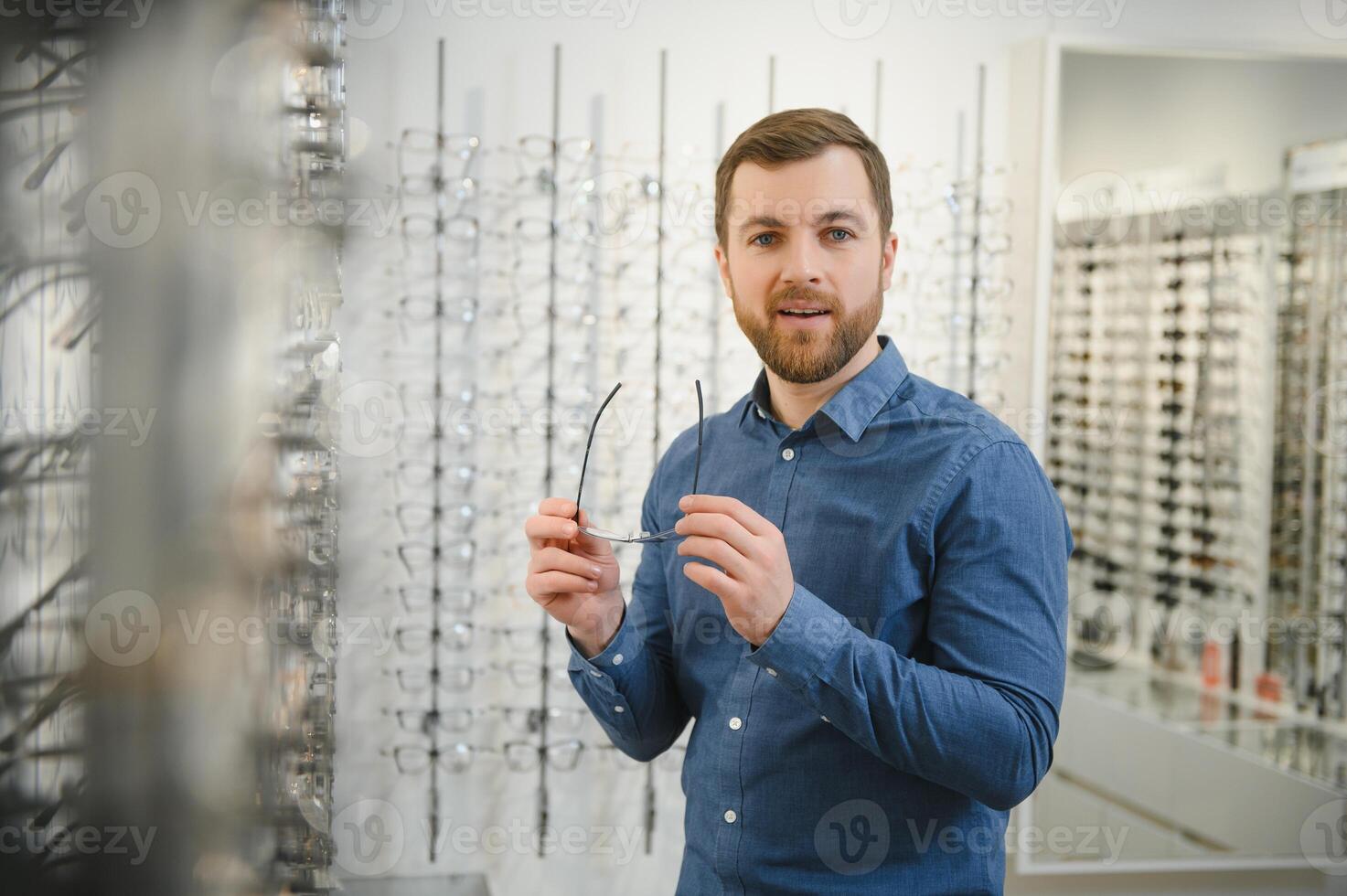 In Optics Shop. Portrait of male client holding and wearing different spectacles, choosing and trying on new glasses at optical store. Man picking frame for vision correction, closeup. photo