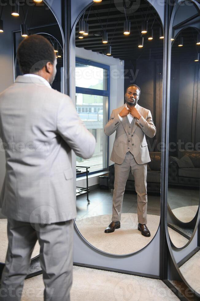 sale, shopping, fashion, style and people concept - african man choosing and trying jacket or suit on and looking to mirror in mall or clothing store photo