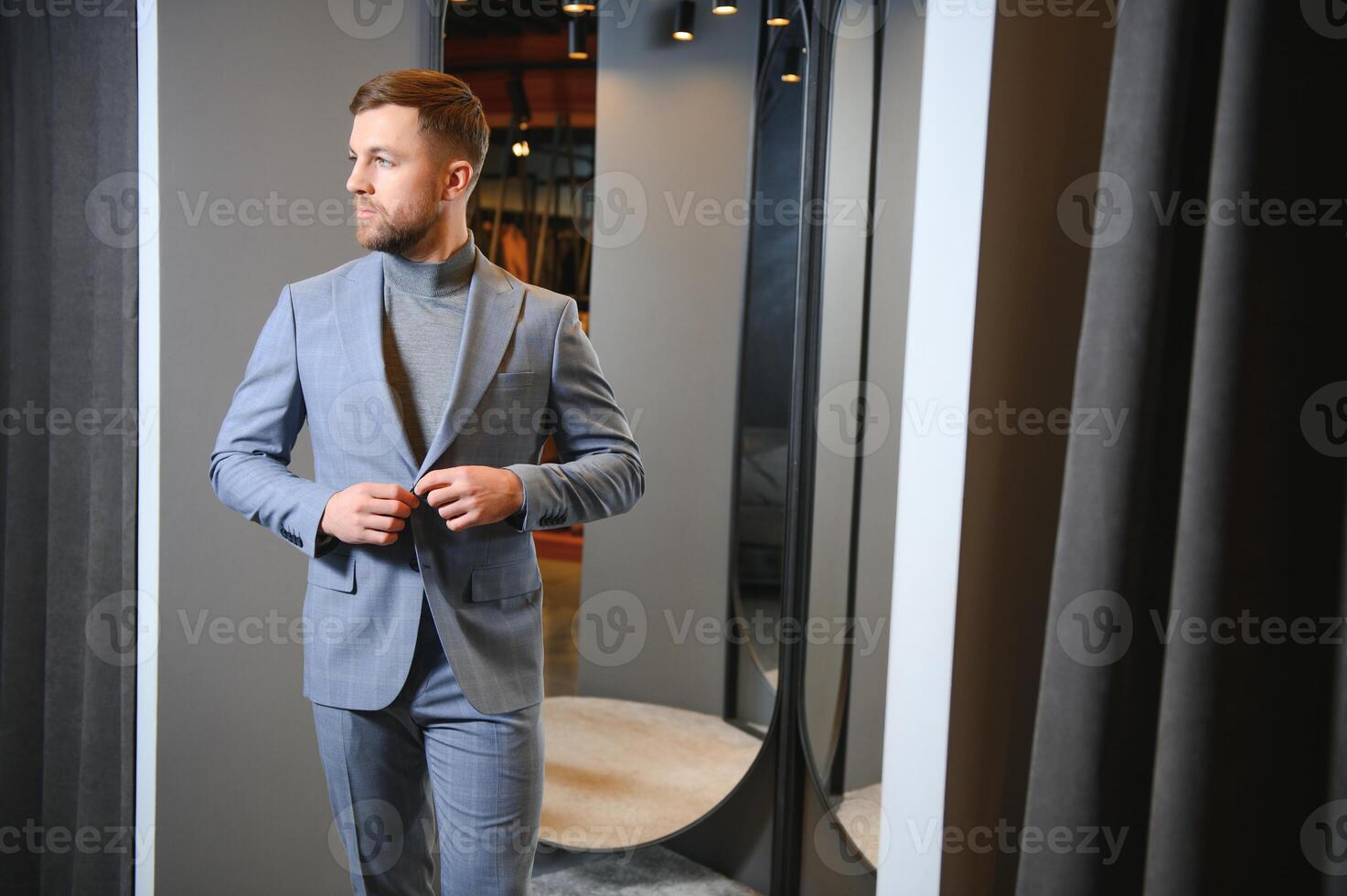 Modern young handsome businessman dressed in classical suit adjusting a tie in front of the mirror while standing in the suit shop photo