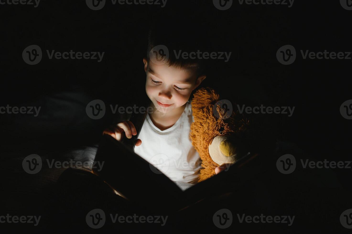 un chico de 5-6 años antiguo es leyendo un libro en el noche en el oscuro debajo un cobija con un juguete oso. foto