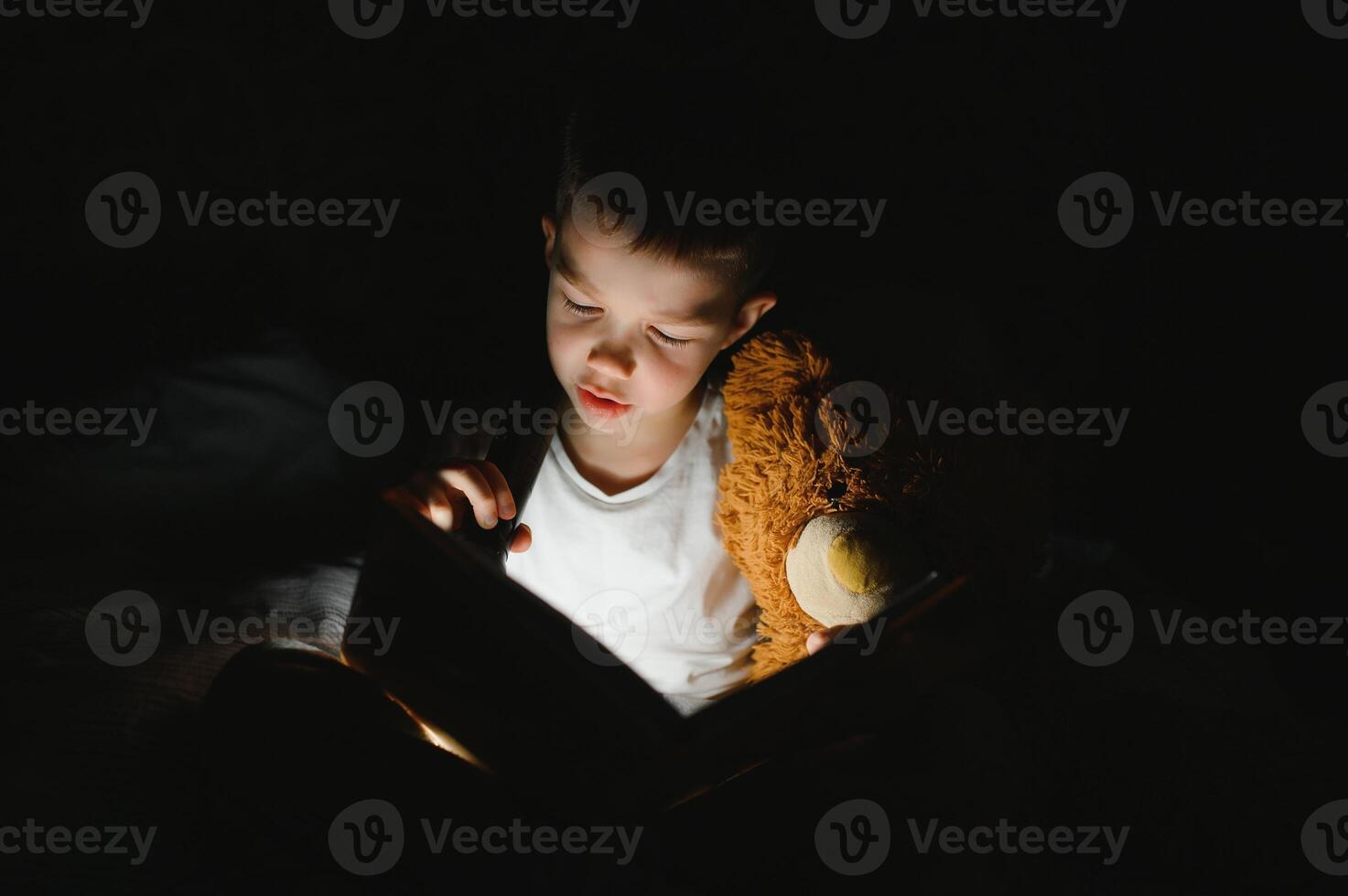 Portrait of cute little boy reading in bed with flashlight in dark room, enjoying fairytales. photo
