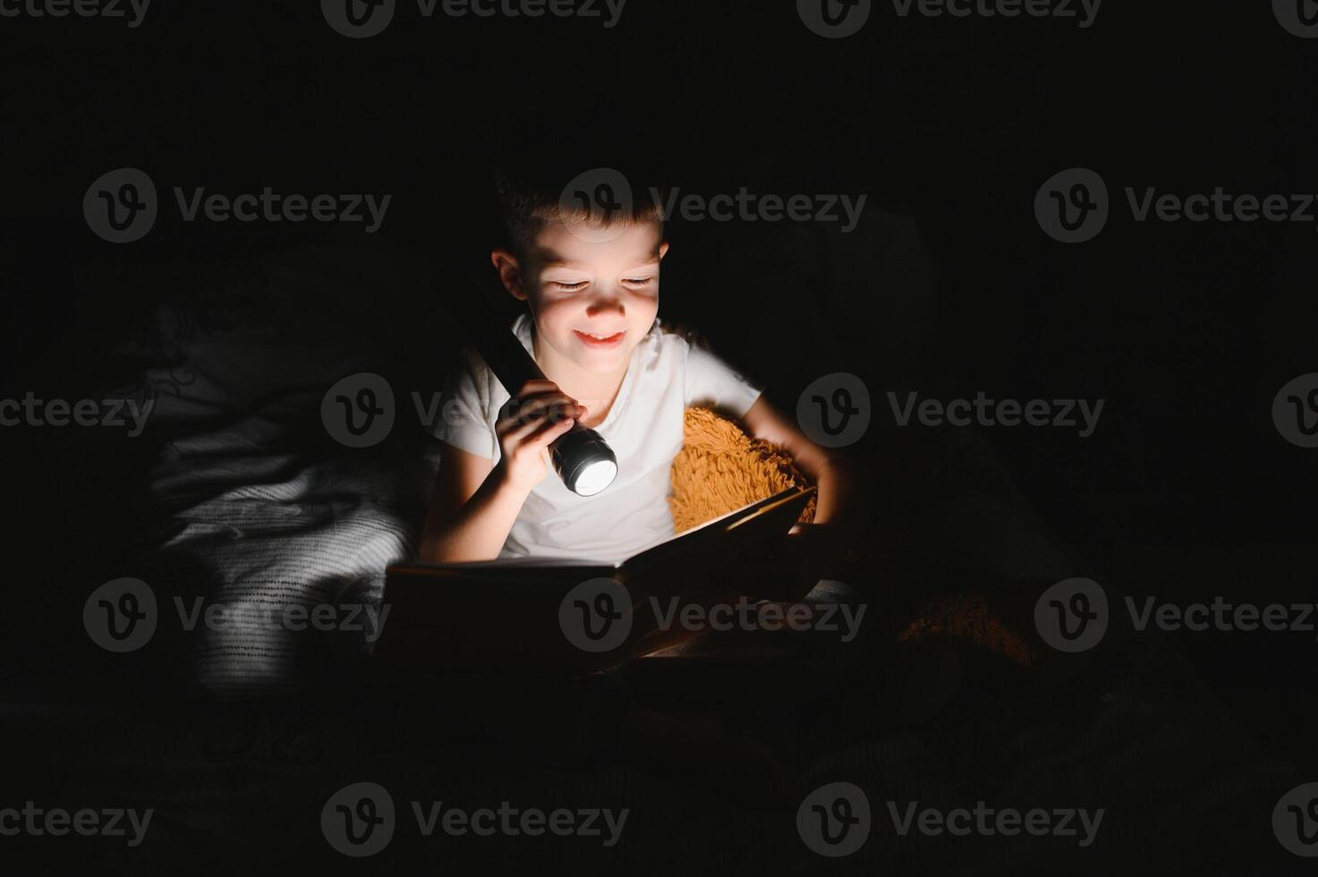 Boy with flashlight reading book under blanket at home. photo