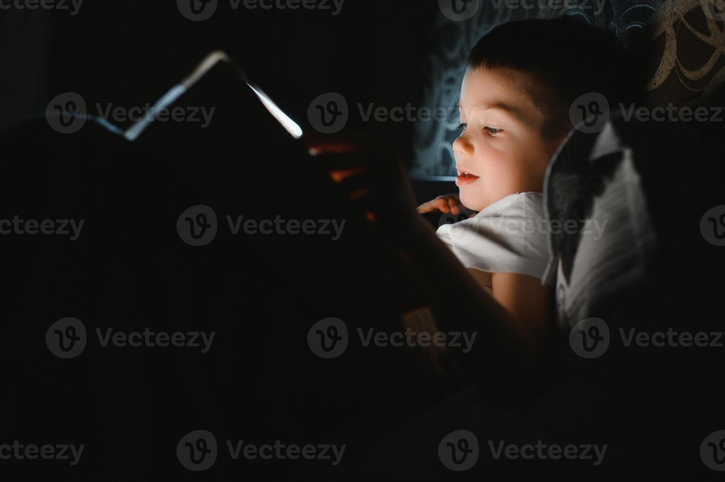 Portrait of cute little boy reading in bed with flashlight in dark room, enjoying fairytales photo