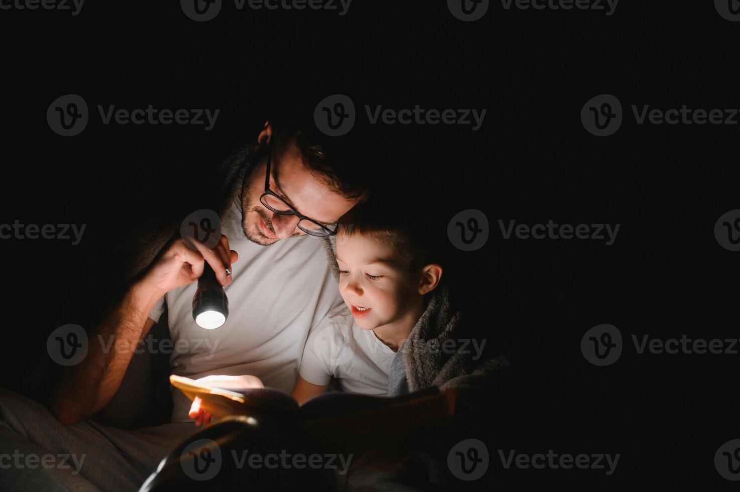 Father and son with flashlight reading book under blanket at home. photo