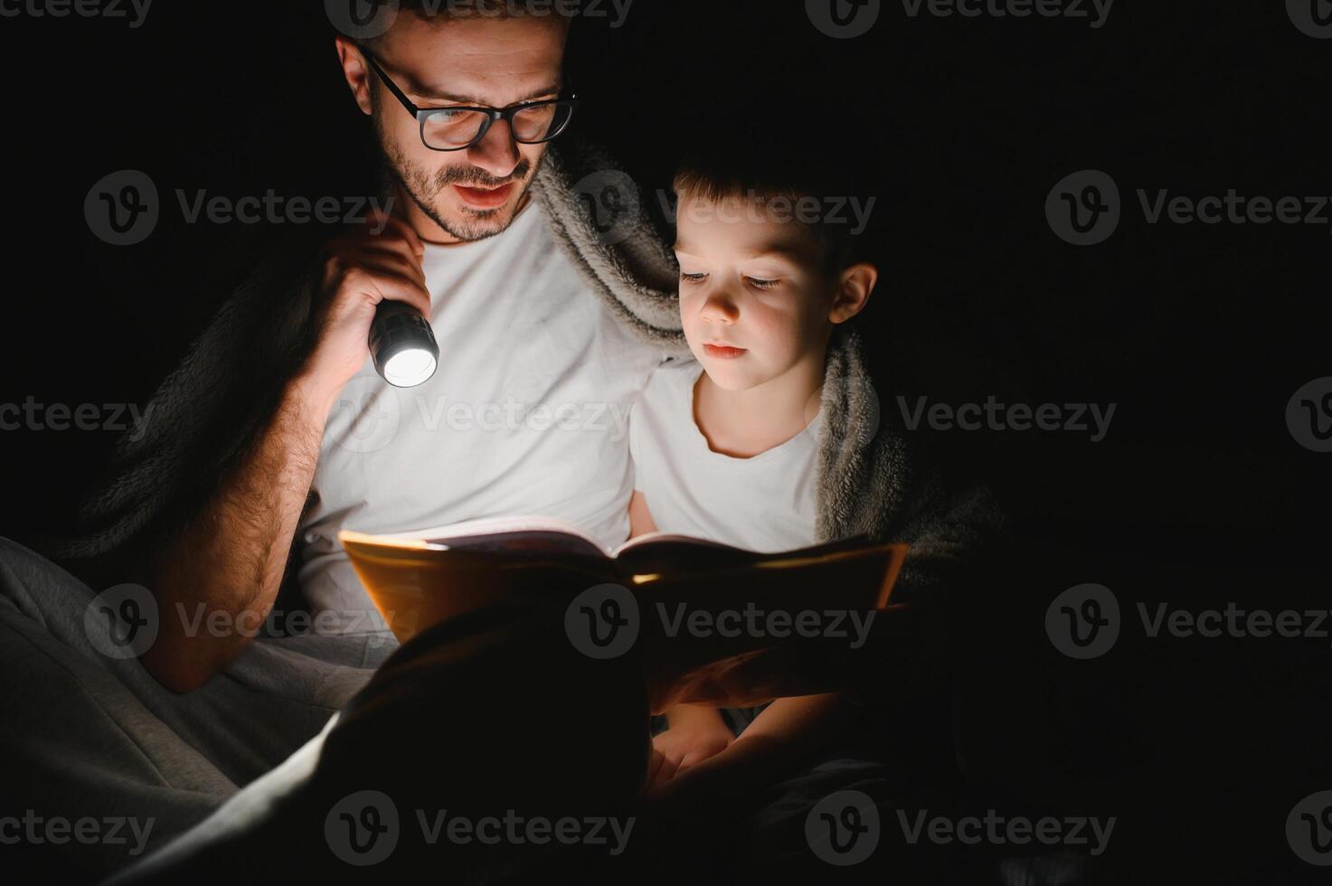 Father and son with flashlight reading book under blanket at home photo