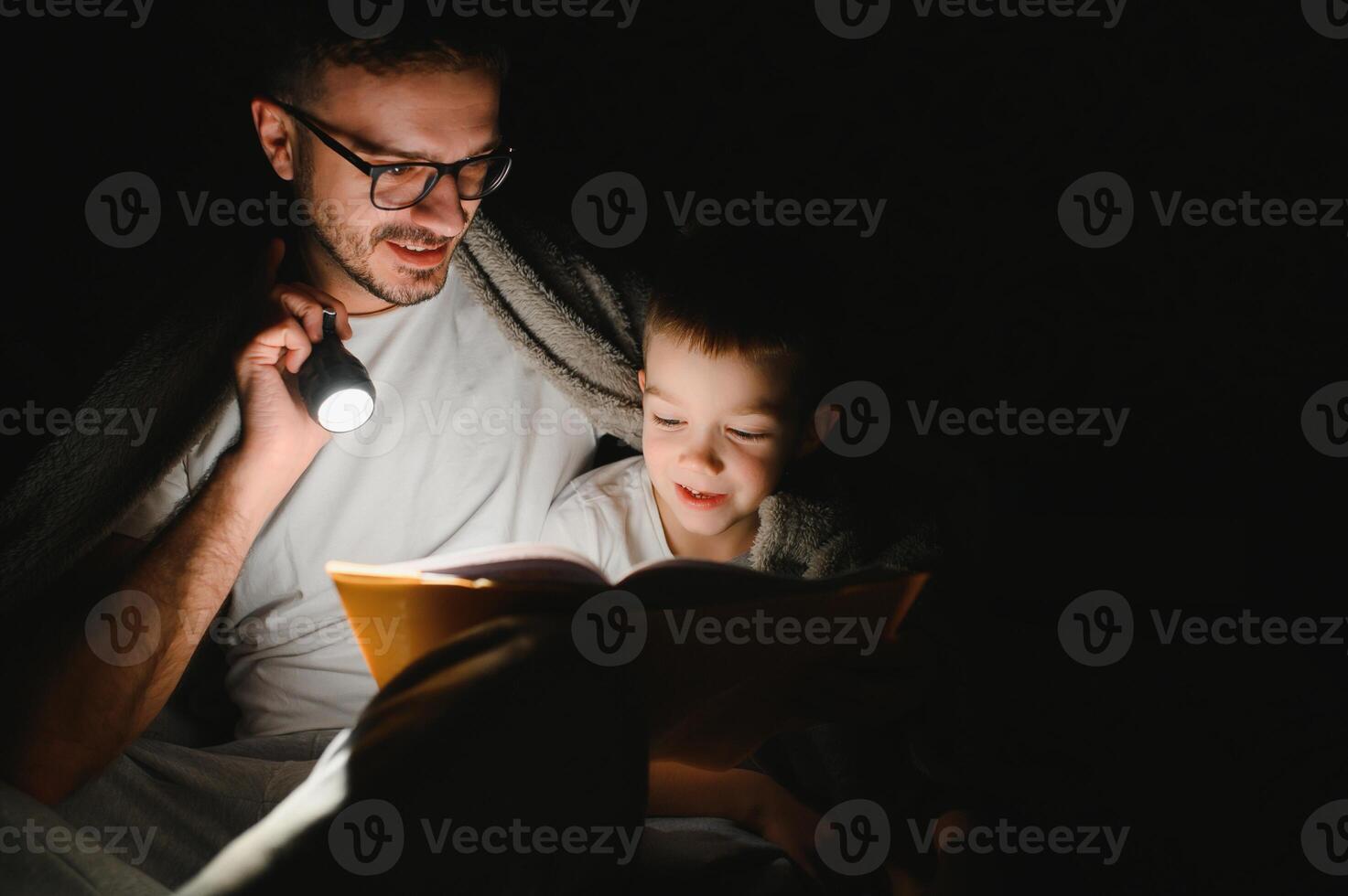 Happy family reading bedtime story under blanket in evening. Father and son spend time together. Father's Day photo