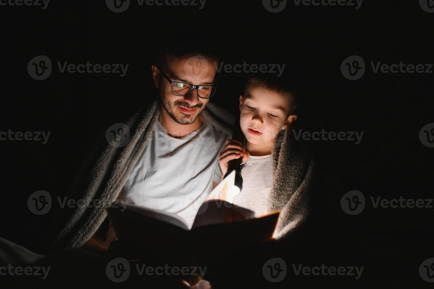 Father and son with flashlight reading book under blanket at home. photo