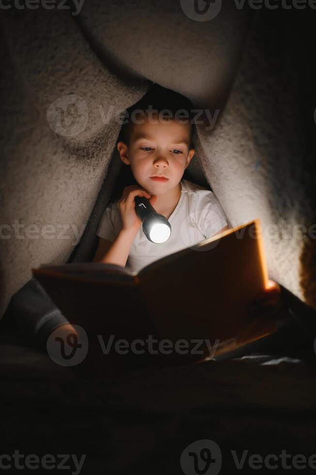 Boy with flashlight reading book under blanket at home. photo