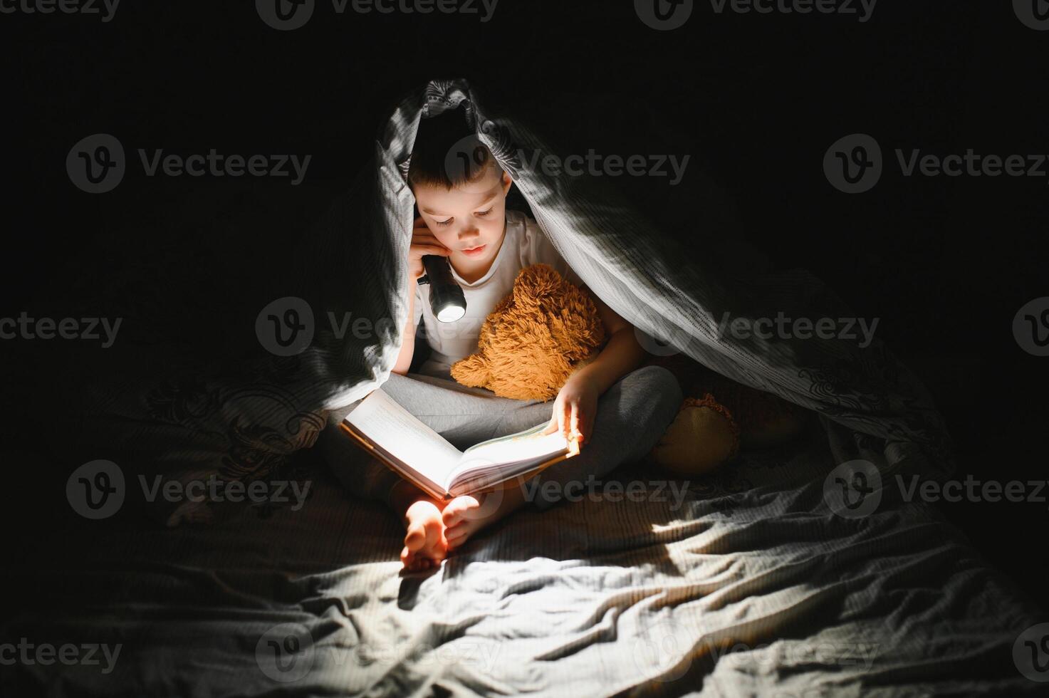 un chico de 5-6 años antiguo es leyendo un libro en el noche en el oscuro debajo un cobija con un juguete oso. foto