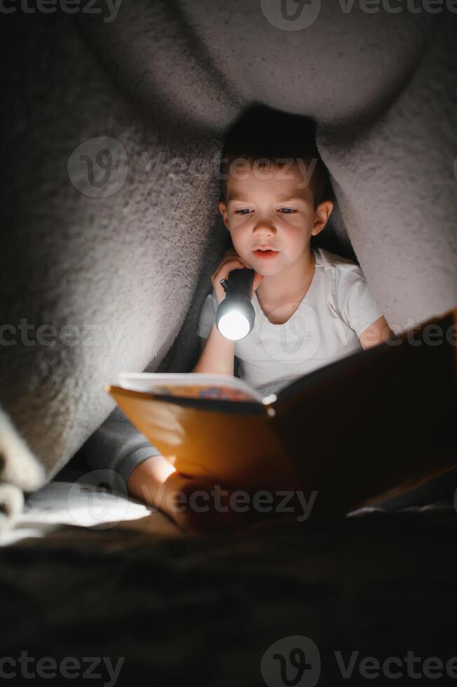 Boy with flashlight reading book under blanket at home. photo