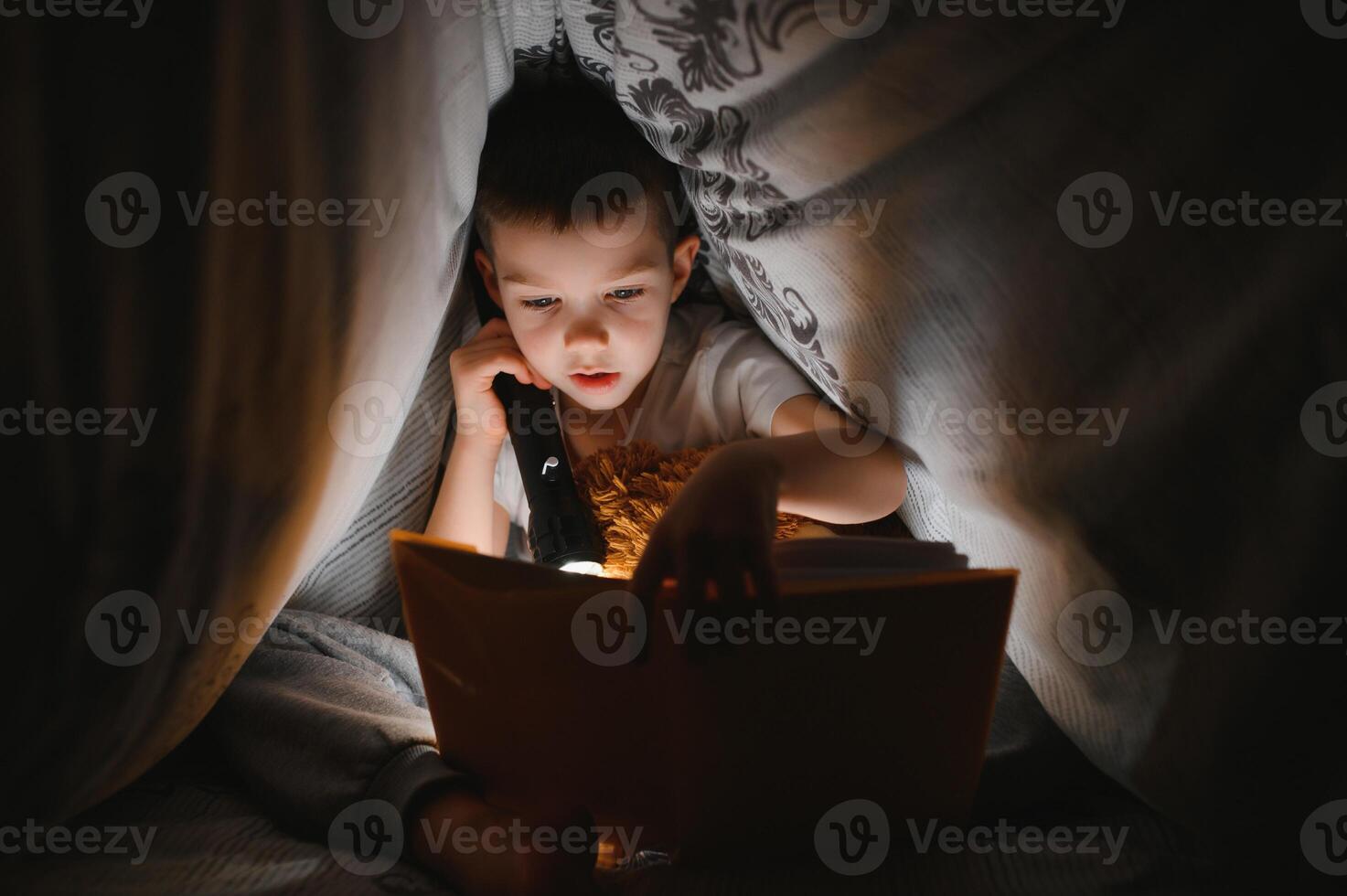 chico con Linterna leyendo libro debajo cobija a hogar. foto