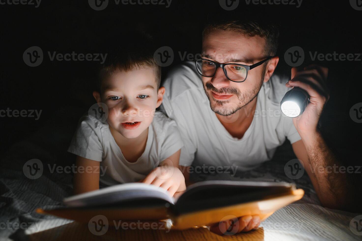 Father and son with flashlight reading book under blanket at home. photo