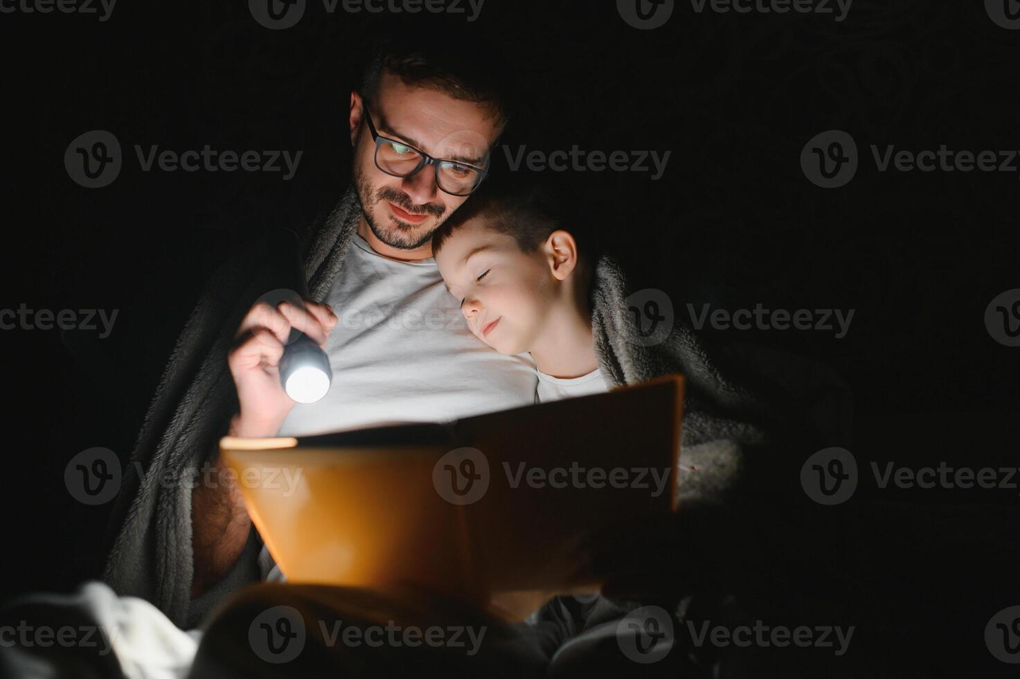 Happy family reading bedtime story under blanket in evening. Father and son spend time together. Father's Day photo