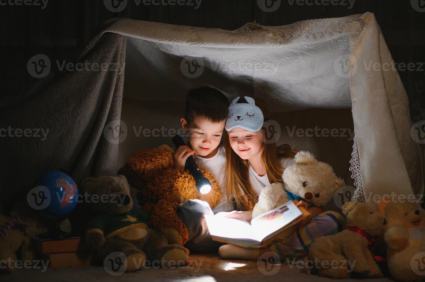 Two children with flashlight read a book under a blanket as a tent. photo