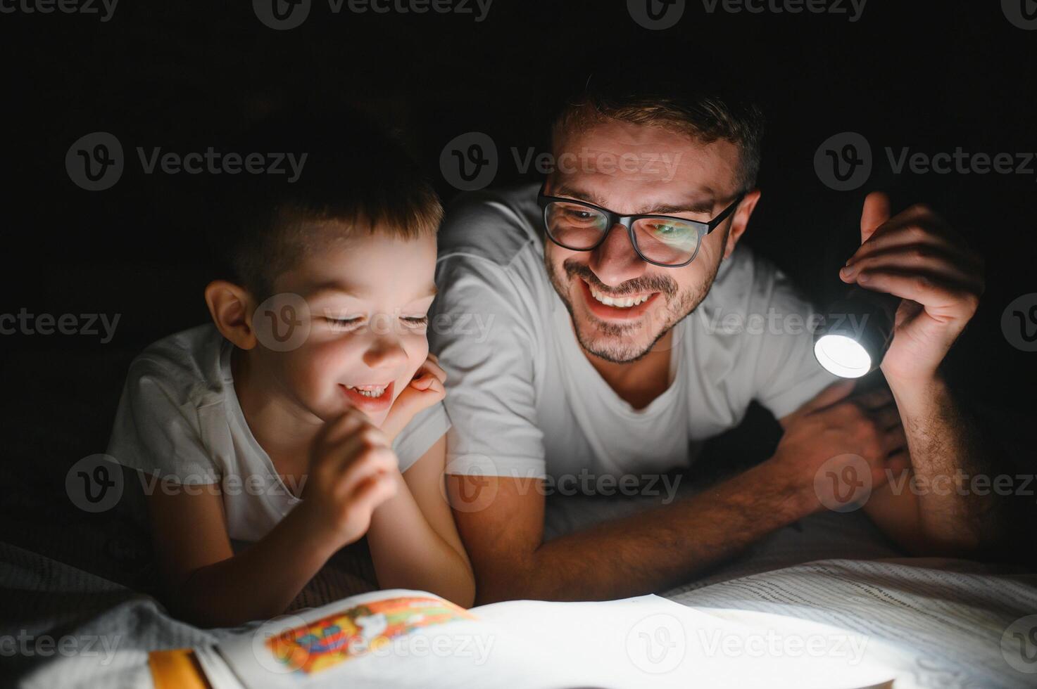 Father and son with flashlight reading book under blanket at home. photo