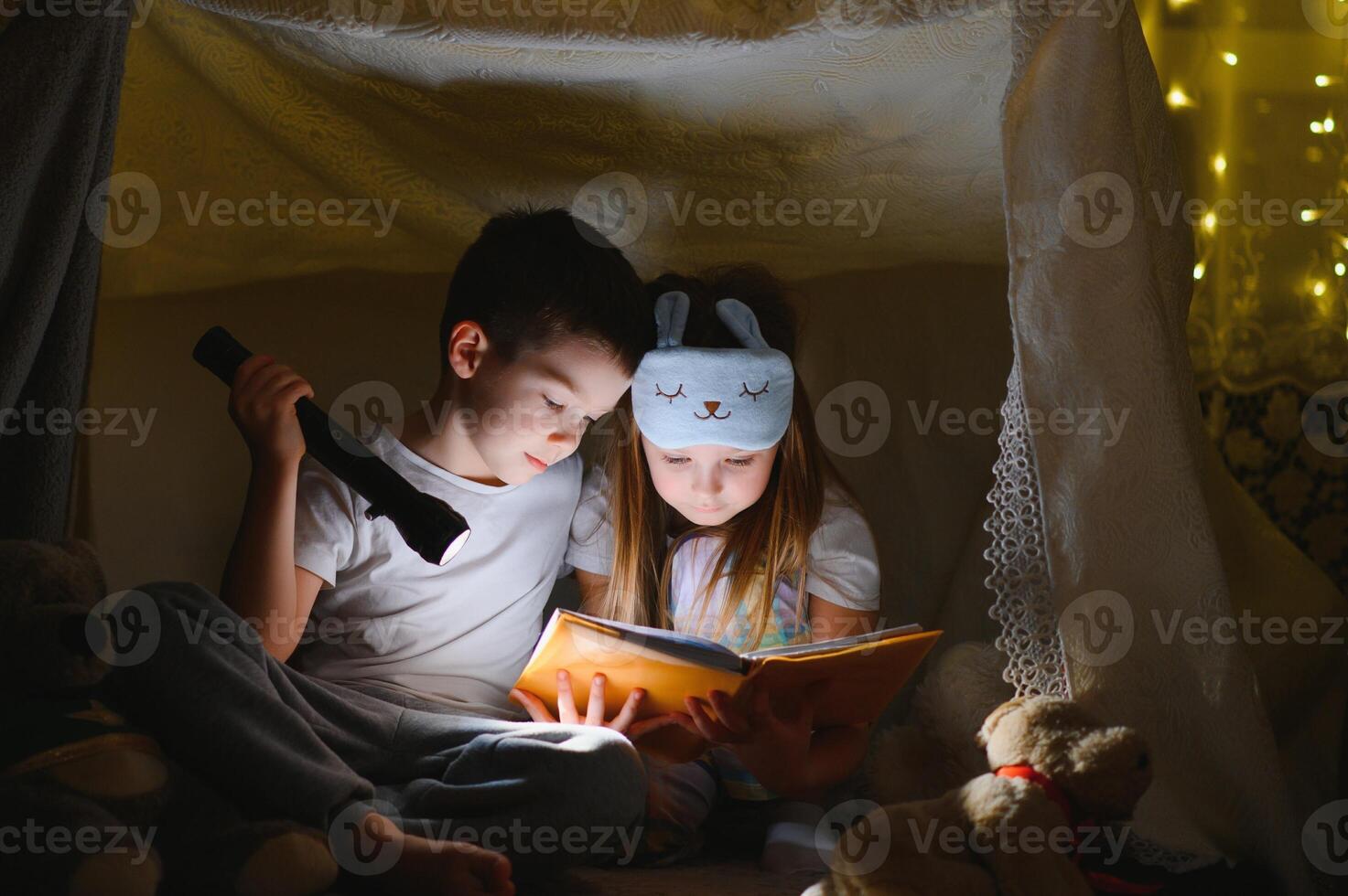 Little kids involving in reading amazing book. They lying in nice toy tent in playroom. Boy holding flashlight in hand. photo