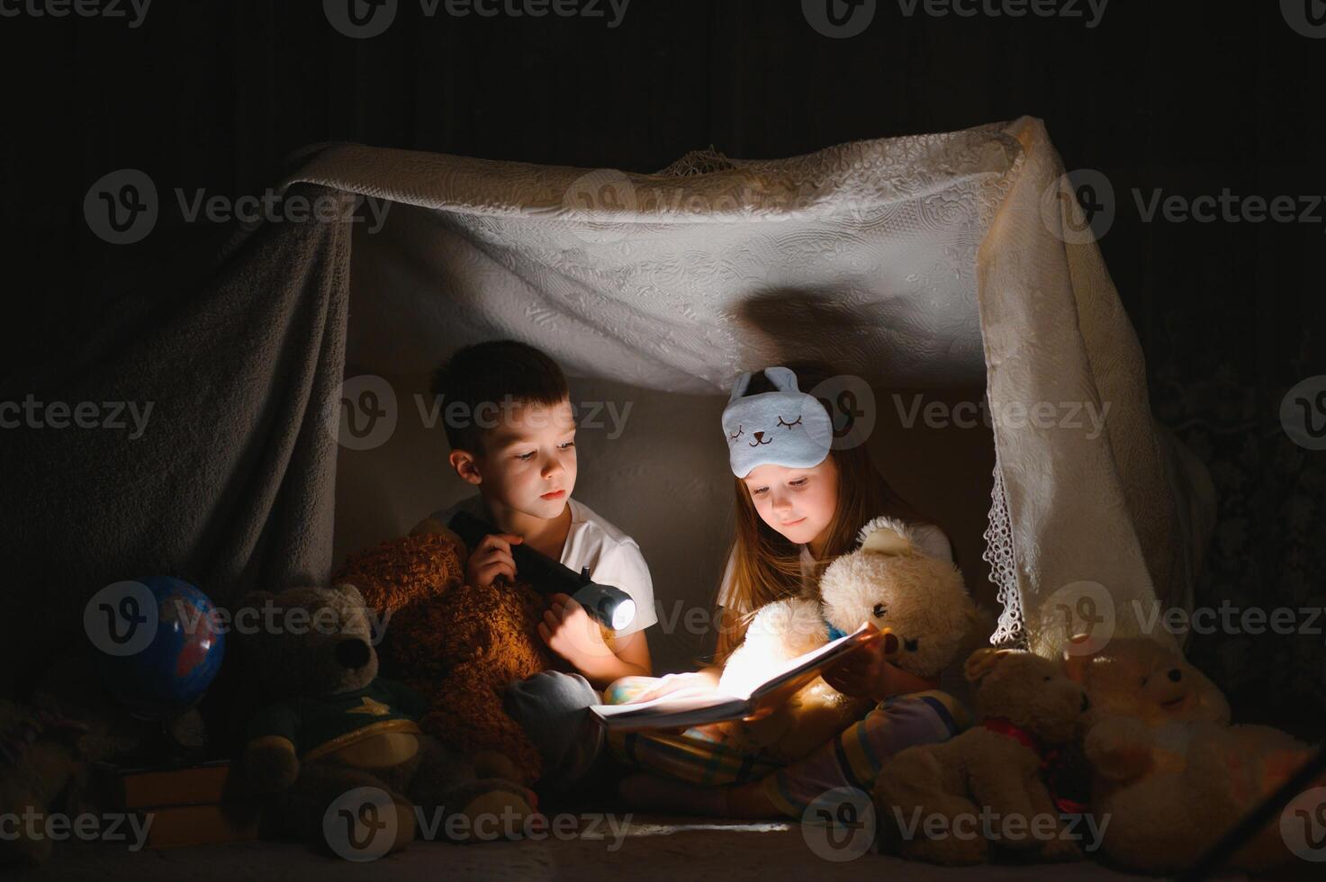 Little kids involving in reading amazing book. They lying in nice toy tent in playroom. Boy holding flashlight in hand. photo