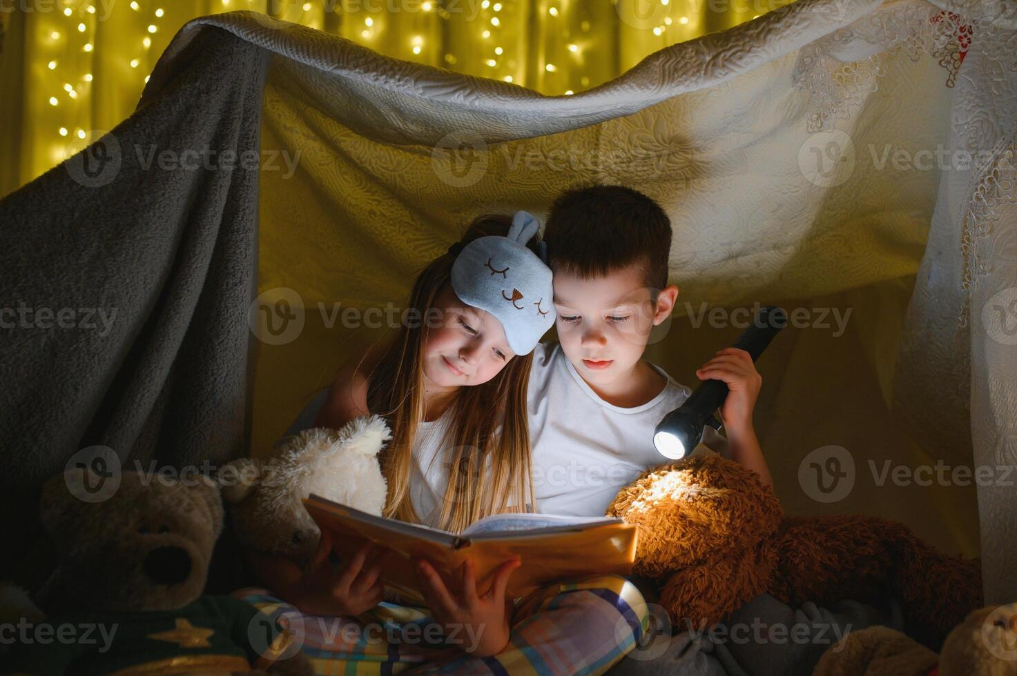 Two children with flashlight read a book under a blanket as a tent. photo