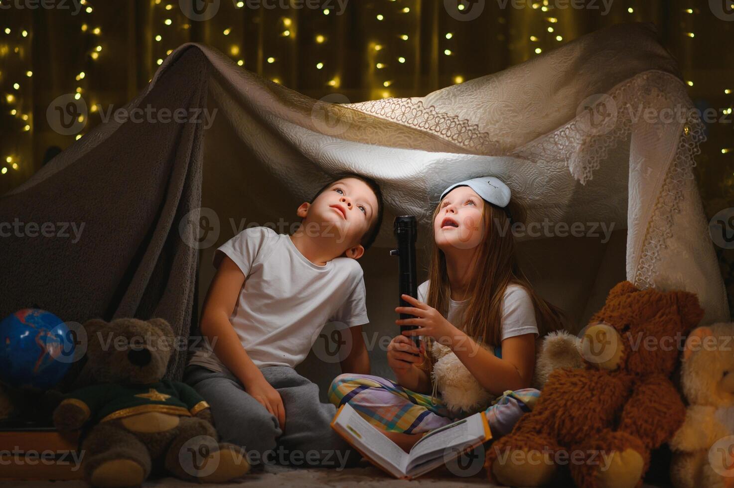dos pequeño niño jugar a hogar en el noche a construir un cámping tienda a leer libros con un Linterna y dormir adentro. concepto de juego, magia, creatividad, alarma sistemas foto
