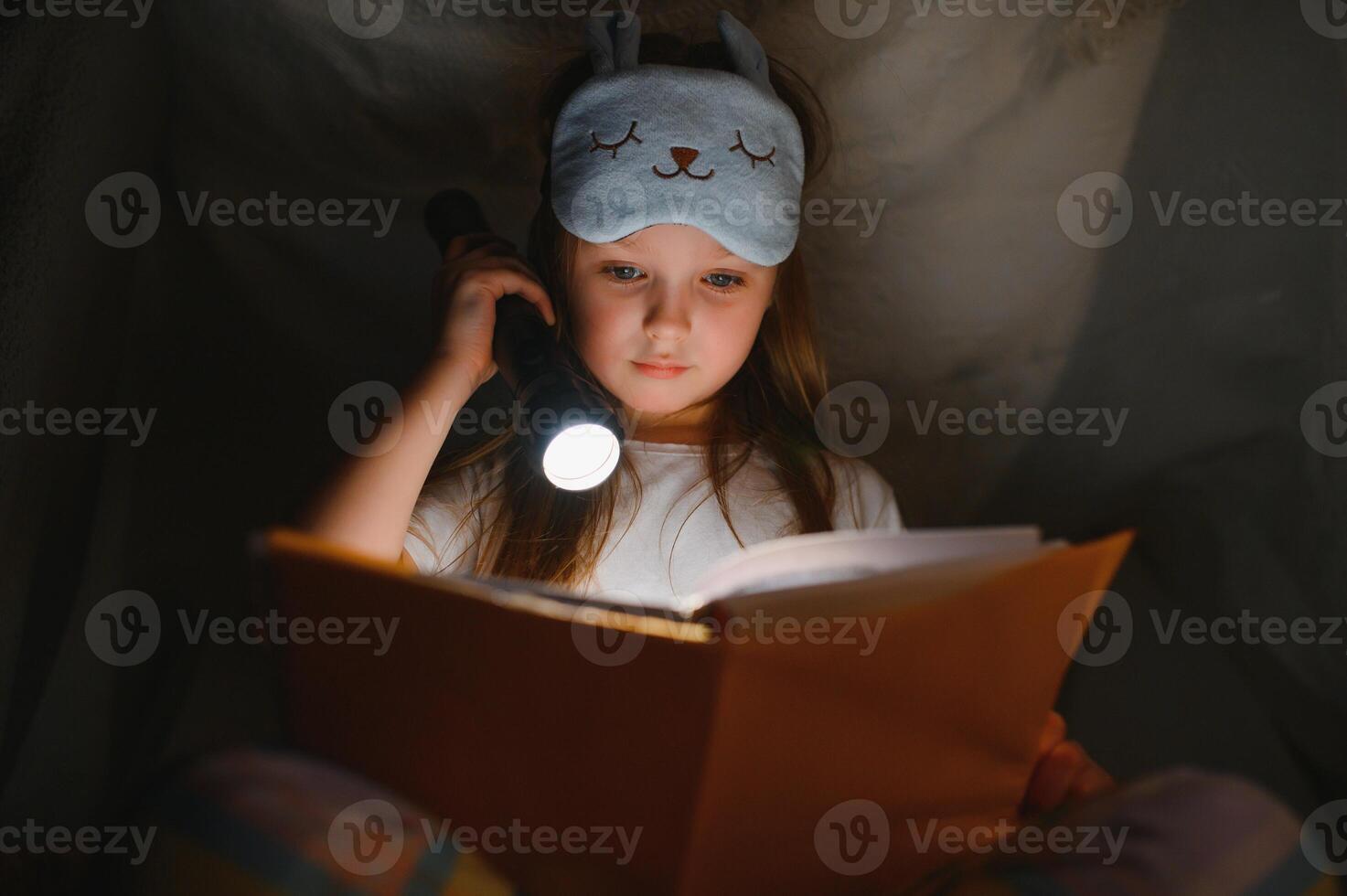 child girl reading with book and flashlight and teddy bear in tent. before going to bed. photo
