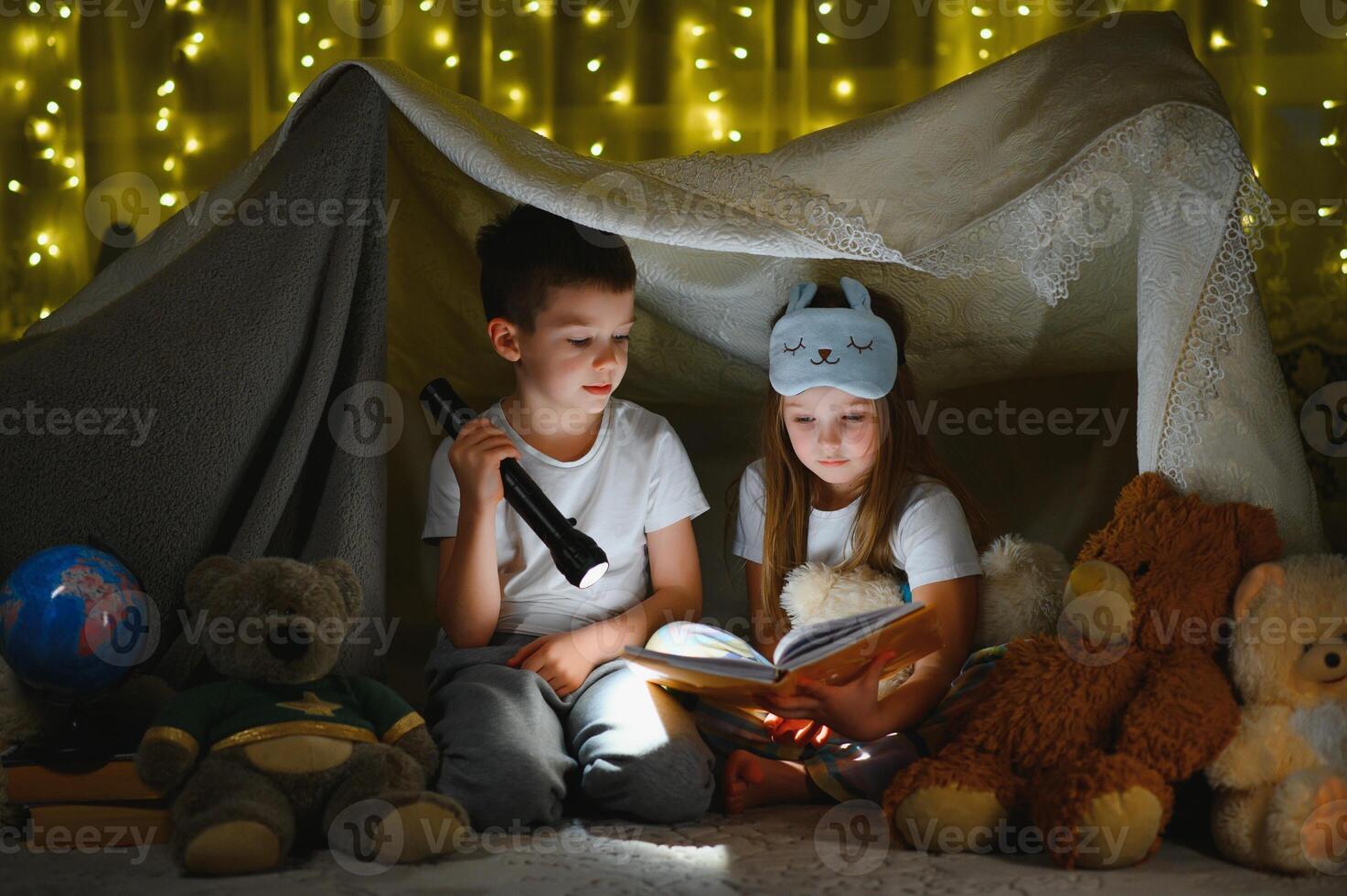 Two children with flashlight read a book under a blanket as a tent. photo