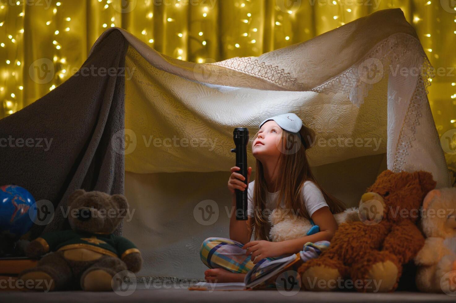 niño niña leyendo con libro y Linterna y osito de peluche oso en carpa. antes de yendo a cama. foto
