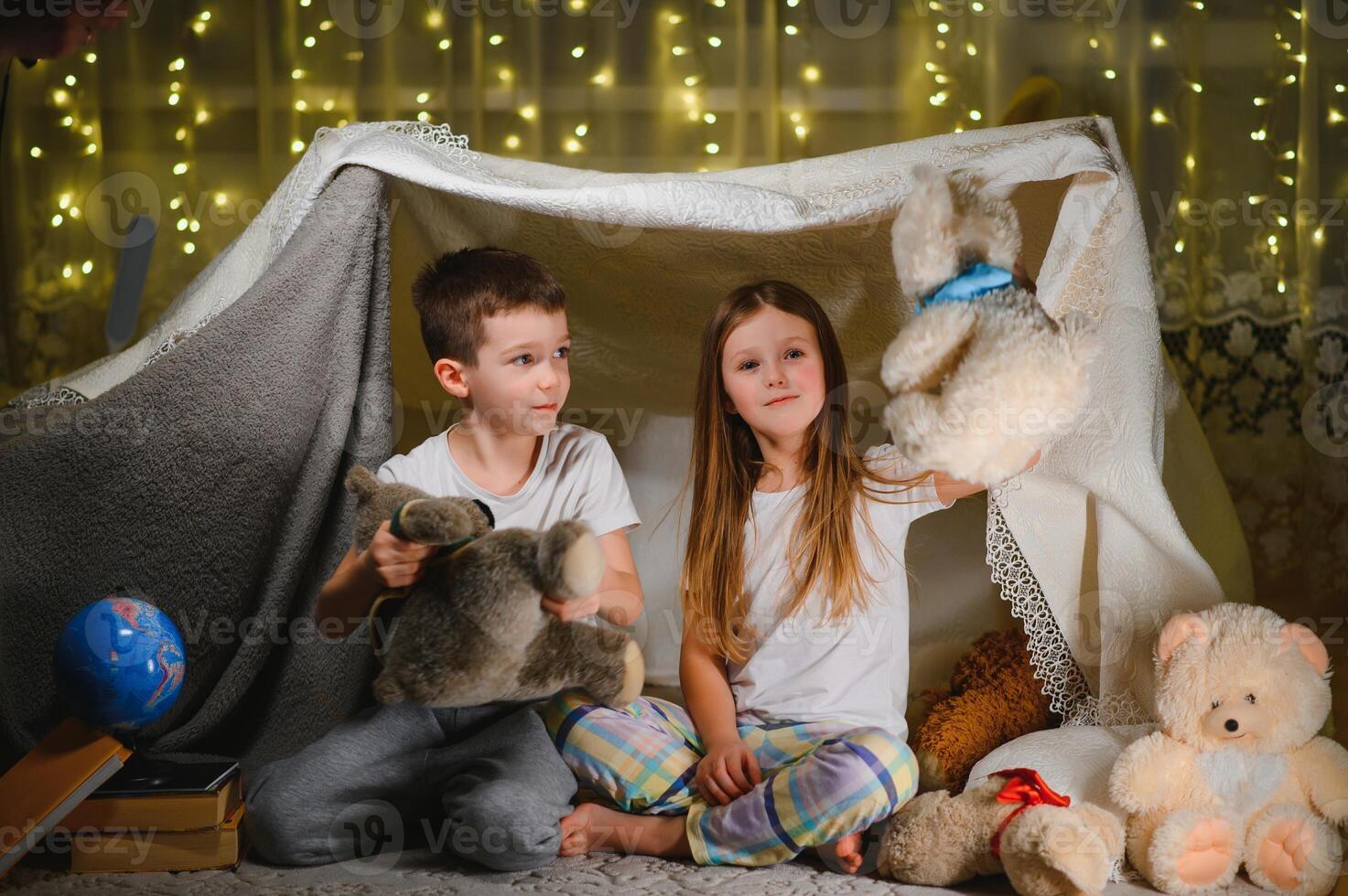 dos pequeño niños jugar a hogar en el noche a construir un cámping tienda a leer libros con un Linterna y dormir adentro. concepto de juego, magia, creatividad, alarma sistemas foto