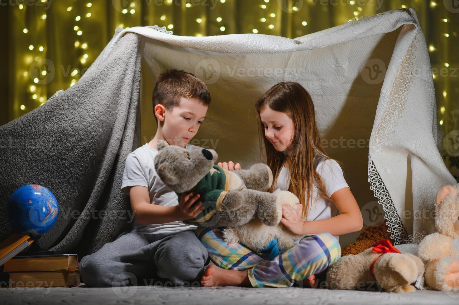 dos pequeño niños jugar a hogar en el noche a construir un cámping tienda a leer libros con un Linterna y dormir adentro. concepto de juego, magia, creatividad, alarma sistemas foto