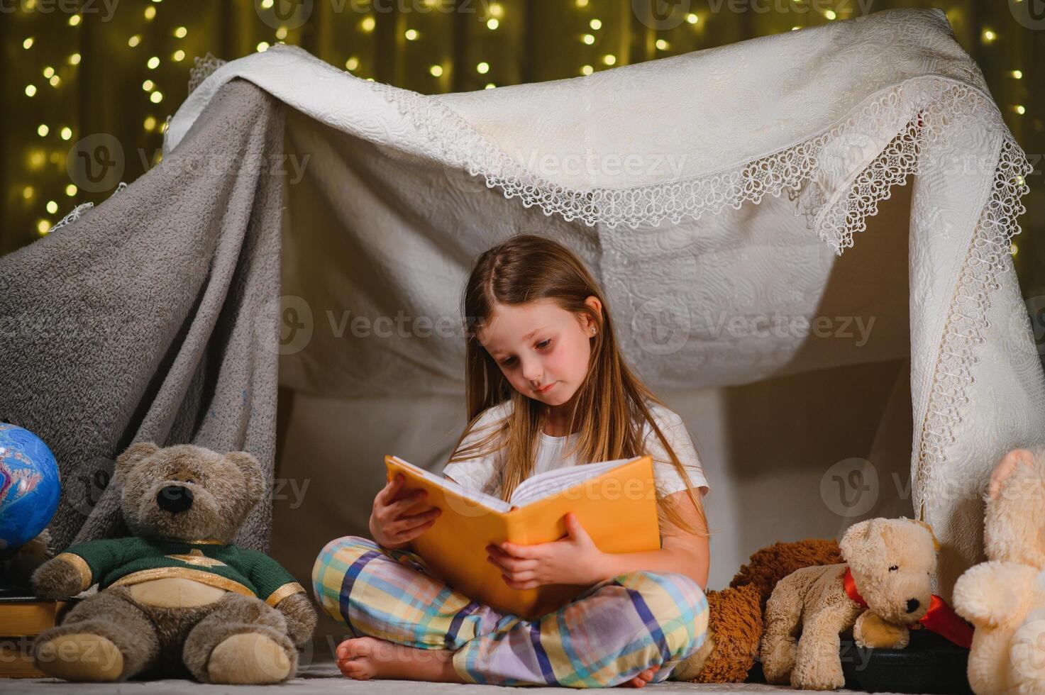 child girl reading with book and flashlight and teddy bear in tent. before going to bed. photo