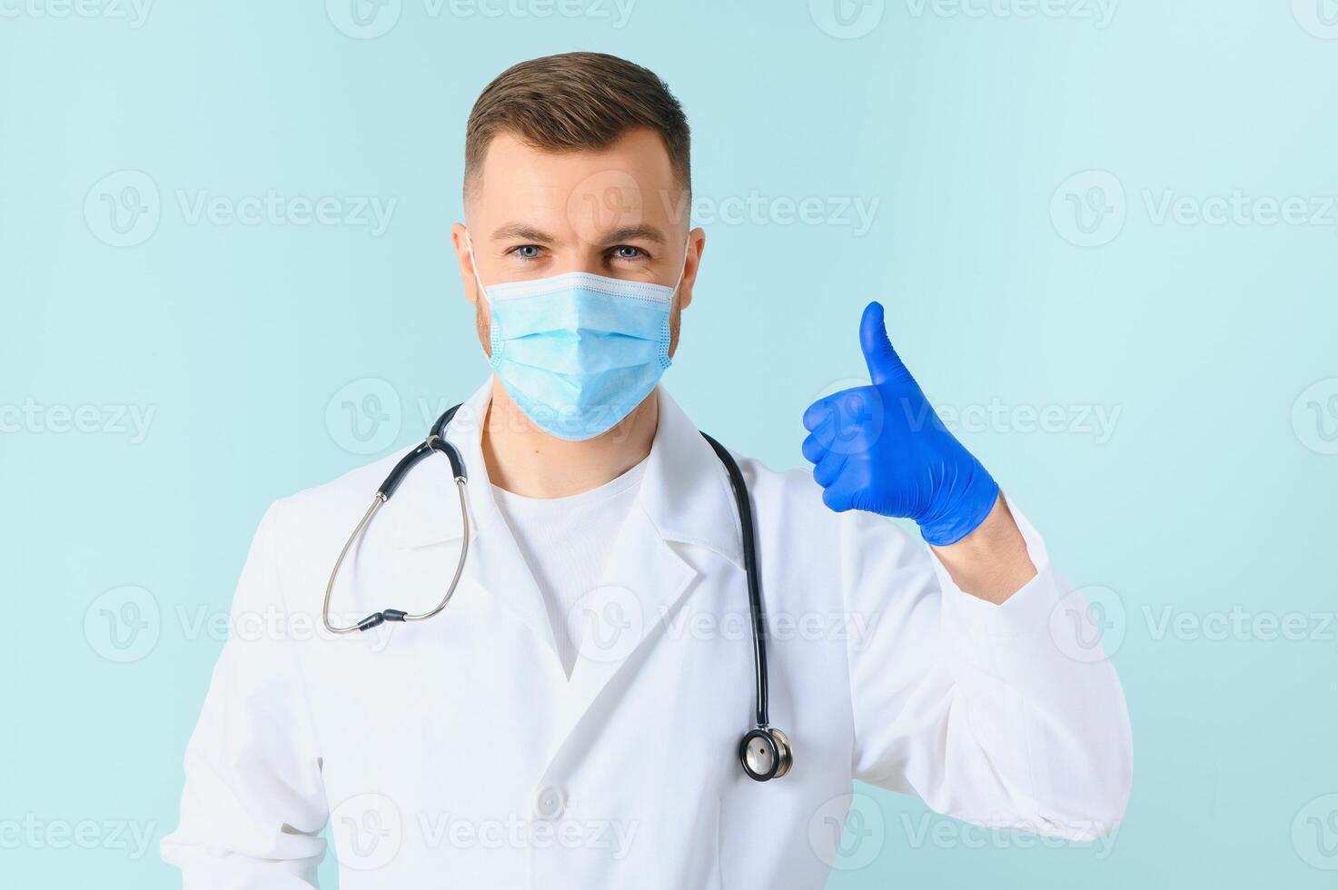 Young male doctor wearing medical mask, and stethoscope round his neck, isolated on blue background photo