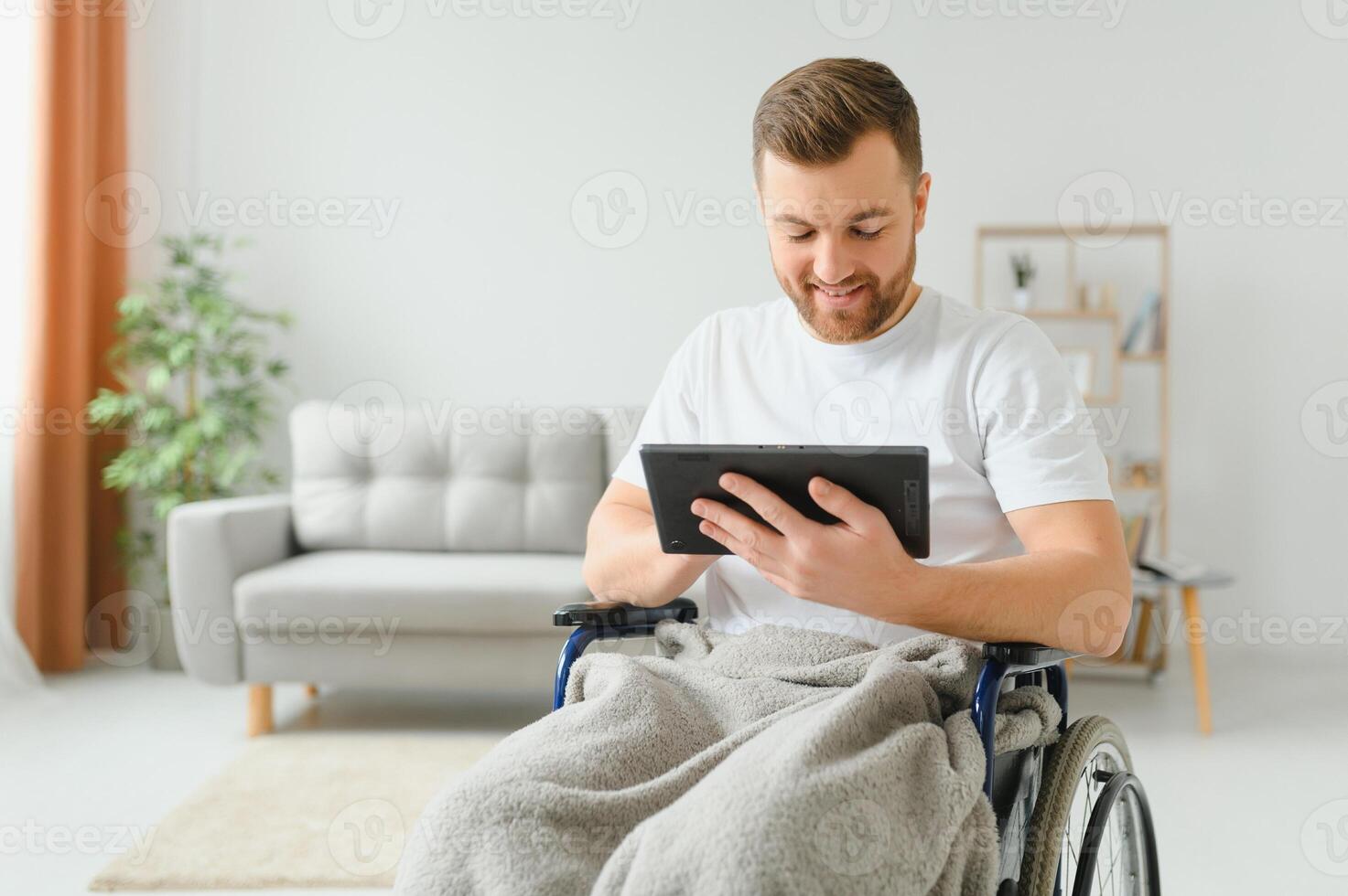 Man in wheelchair using tablet computer at home. Positive retired male with physical disability browsing web on touch pad, watching video online indoors. photo