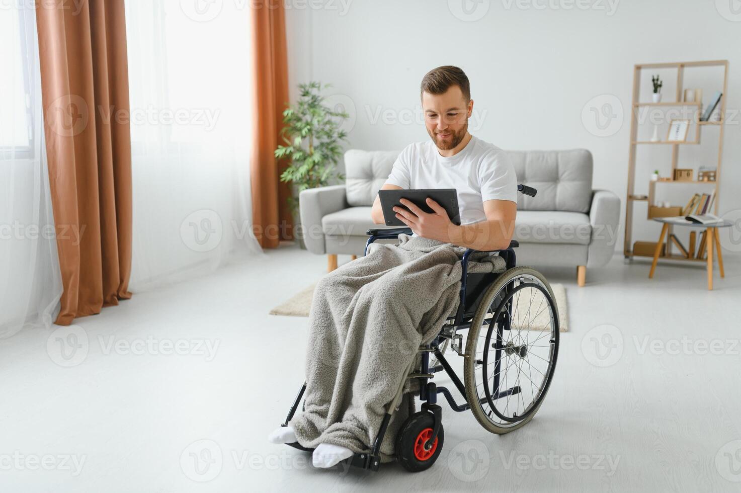 Man in wheelchair using tablet computer at home. Positive retired male with physical disability browsing web on touch pad, watching video online indoors photo