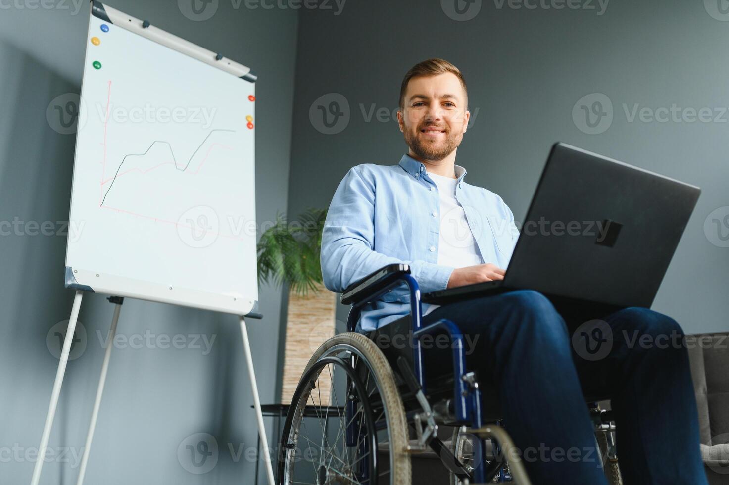 Young man with special needs in casual clothes working on wireless laptop. Male freelancer working from home while sitting in wheelchair. photo