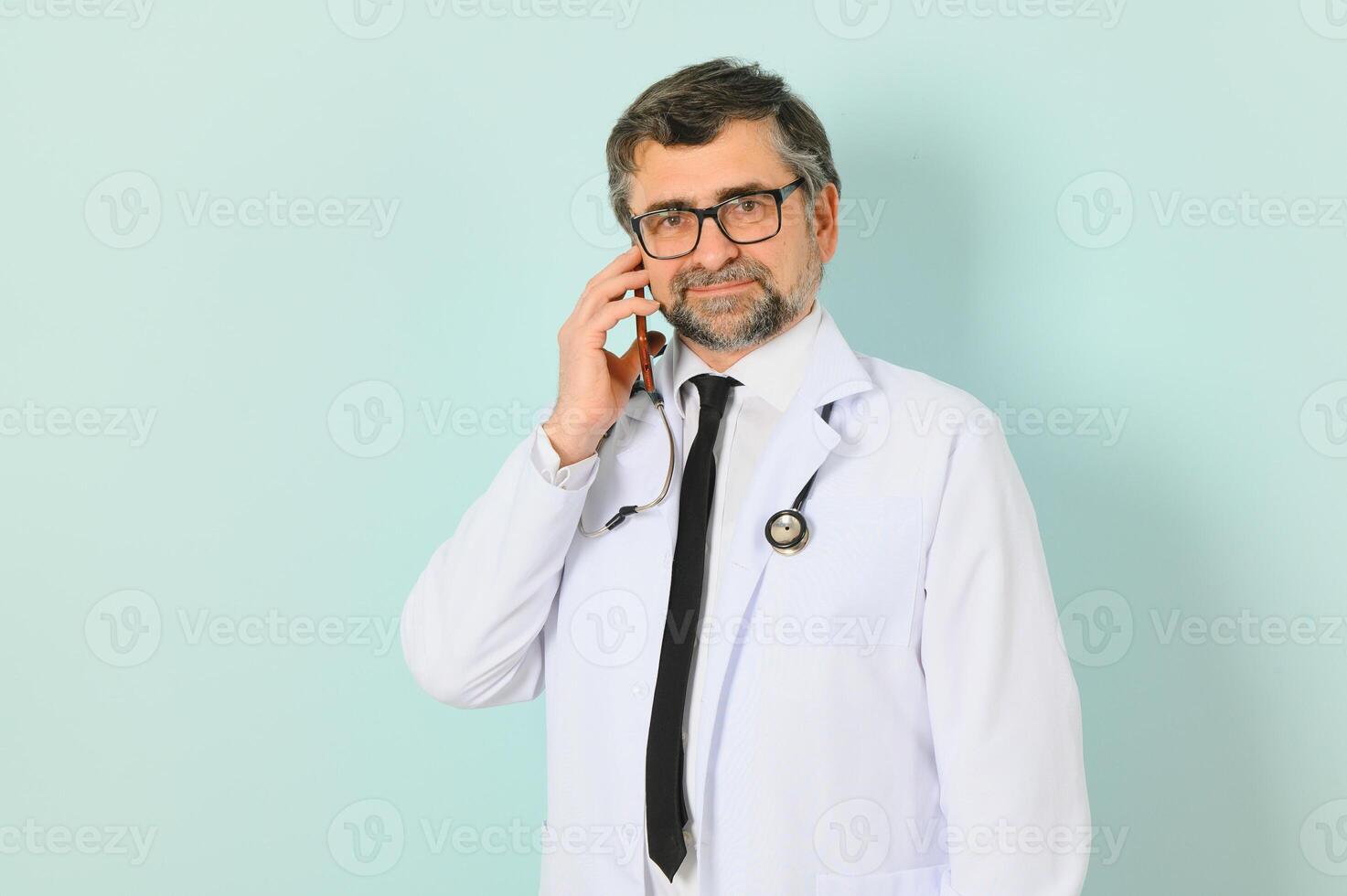 Photo of medical doctor wearing white coat and stethoscope holding mobile phone standing isolated over blue background.