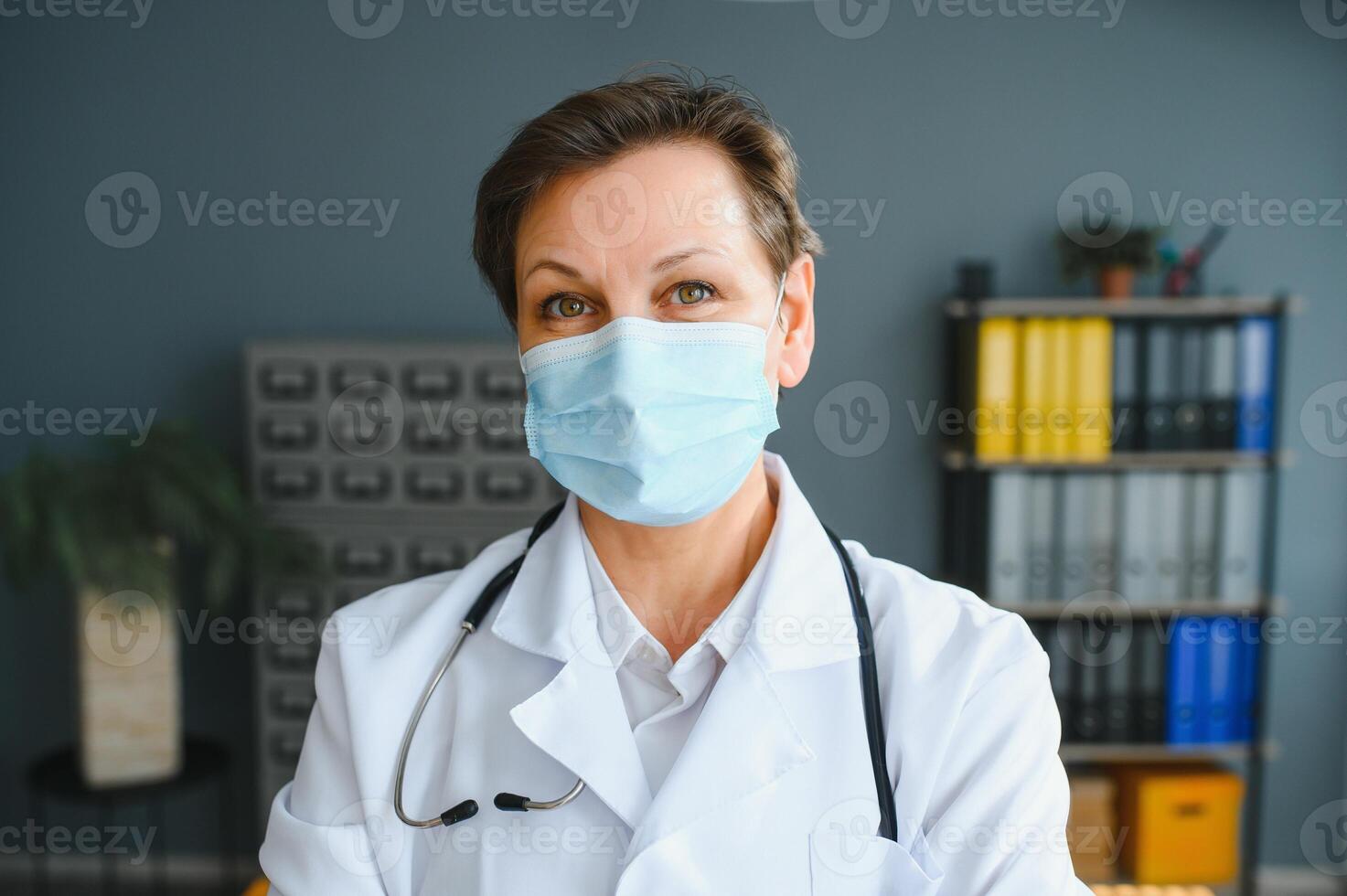 Older female doctor wearing face mask and white medical coat standing in hospital. Portrait photo