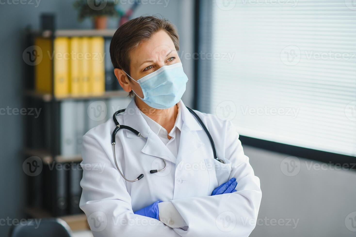 Older female doctor wearing face mask and white medical coat standing in hospital. Portrait photo